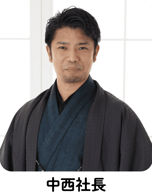 短くウェーブした黒髪に、整えられた髭を生やした男性が、紺色の伝統的な着物と黒い羽織を身に着けている。大きな窓の前に立ち、柔らかな笑みを浮かべ、自然光が顔を照らしている。.