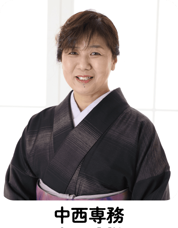 黒と紫の着物を着た黒髪のショートヘアの中年女性が、優しく微笑んでいる。彼女は白い枠の入った明るい窓の前に立っており、背景は柔らかな光に照らされている。.