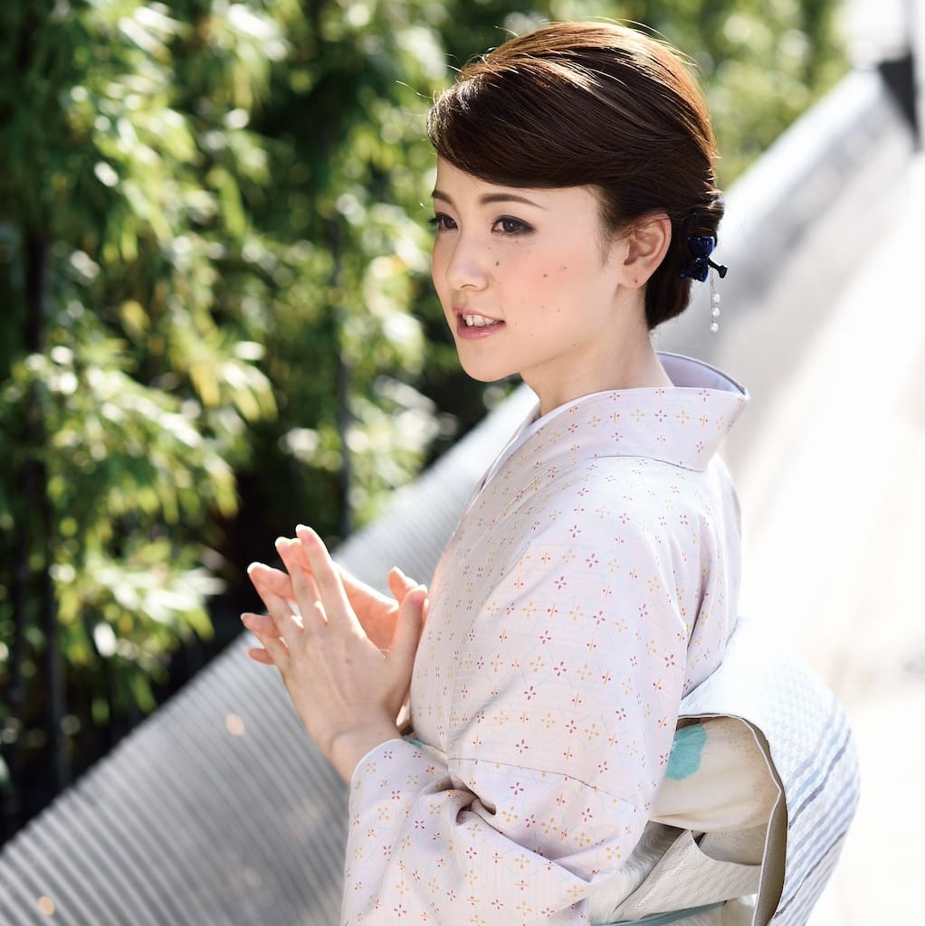 淡い模様の着物を着た女性が、緑の近くの屋外に右を向いて立っている。両手を優しく組み、茶色のショートヘアはきちんとセットされ、青い簪が飾られている。陽光が、彼女の穏やかな表情と伝統的な衣装を際立たせている。.