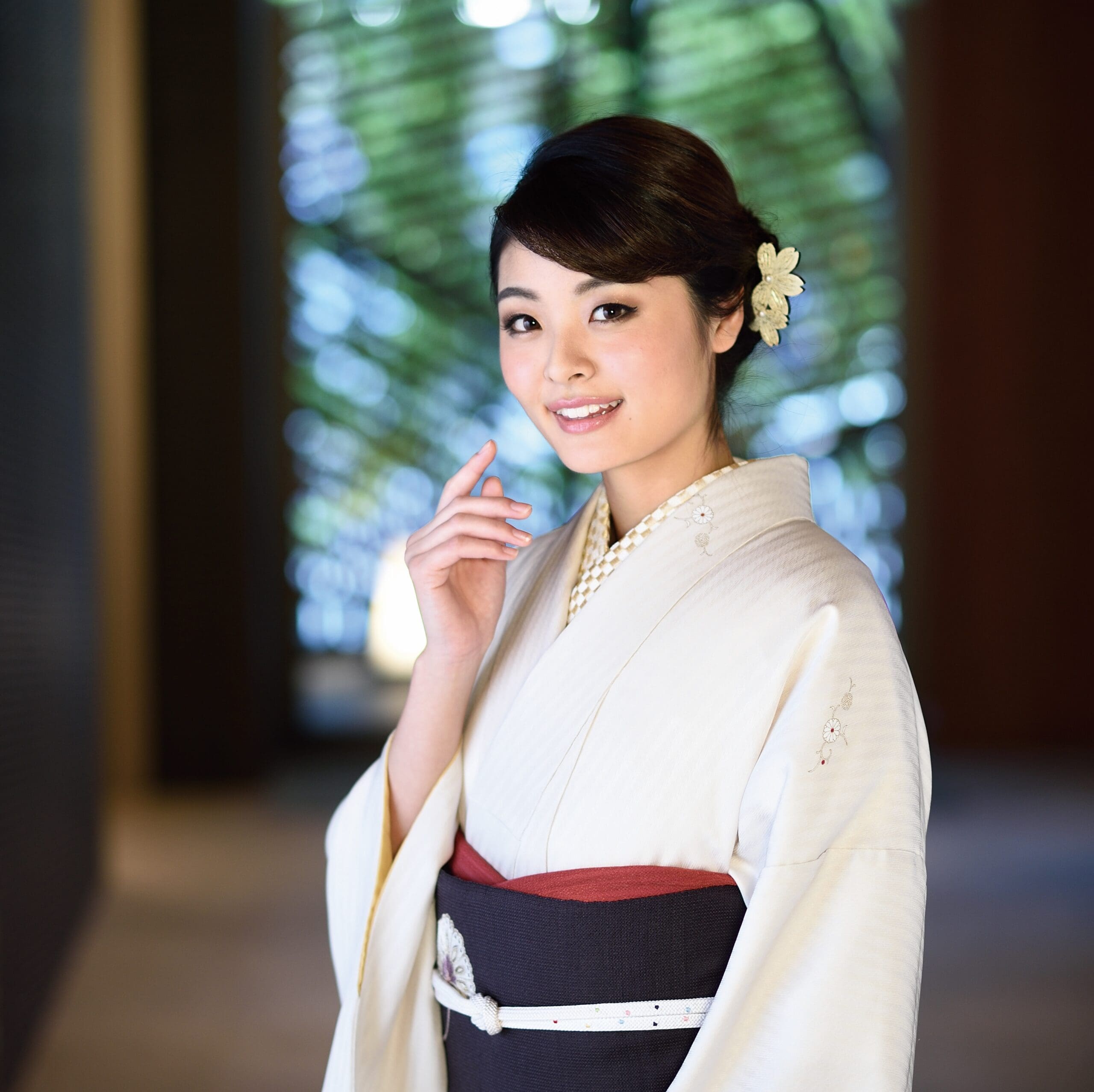 クリーム色の着物に濃い帯を締めた若い女性が、室内に立って優しく微笑んでいる。淡い花の髪飾りをし、髪はセットされている。背景は緑と茶色のトーンで柔らかくぼかされ、窓と自然光を思わせる。.