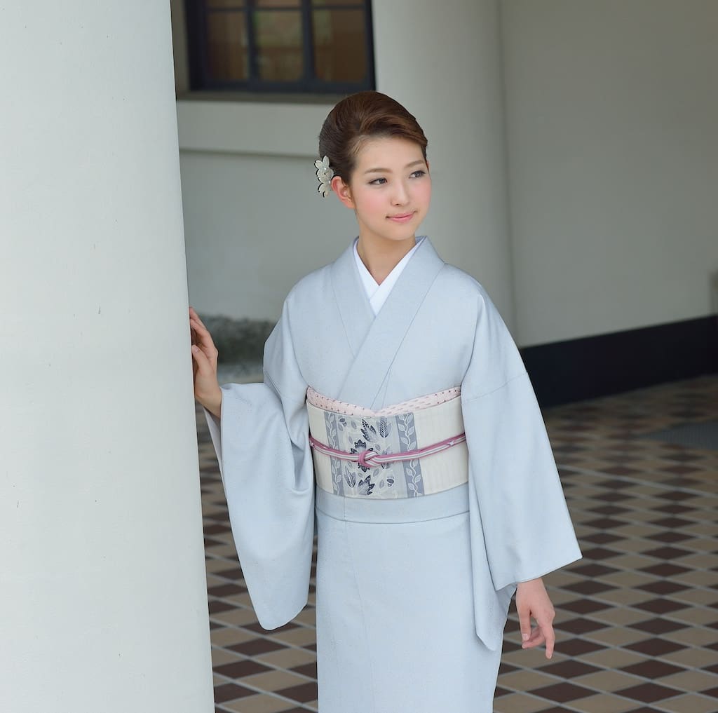 淡いグレーの着物に柄物の帯を締めた若い女性が、室内に立っている。上品なアップヘアに飾り髪を添え、白い柱に軽く触れている。床には市松模様のタイルが敷かれ、彼女はどこか遠くを見つめながら、優しく微笑んでいる。.