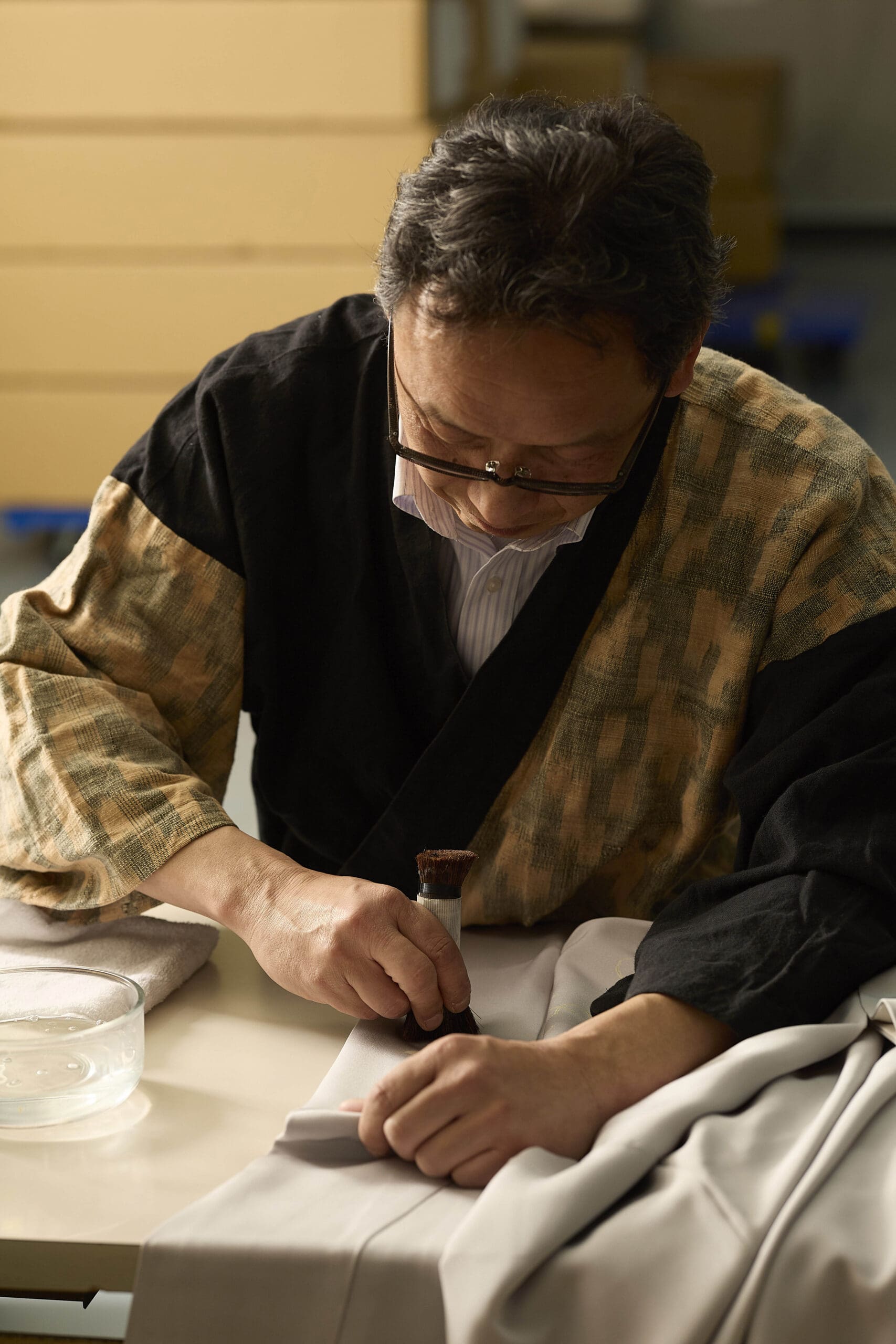 伝統的な和服を着た年配の男性が、テーブルの上で一枚の布に丁寧にスタンプを押している。彼は眼鏡をかけ、集中している。近くには水の入った浅いガラスのボウルが置かれている。背景はやわらかくぼかされ、彼の作業に注意を集中させている。.
