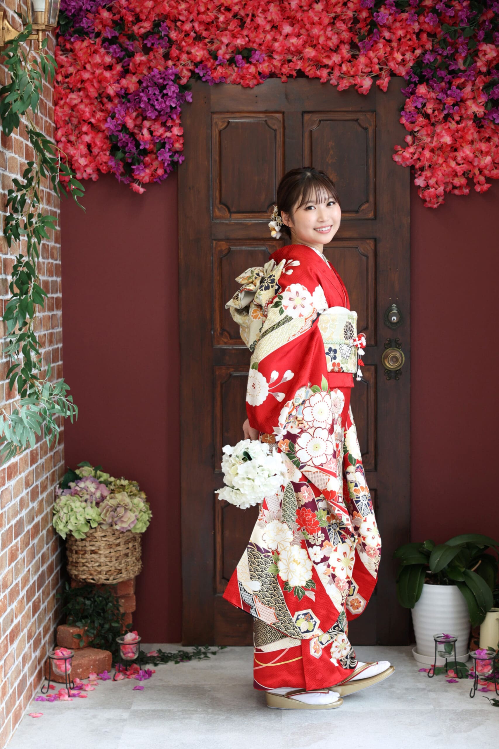 赤い花柄の着物を着た笑顔の女性が白い花を持って横向きに立っている。花のヘアアクセサリーをつけている。彼女の後ろには、鮮やかなピンクと紫の花で縁取られた木製のドアがある。植物と編み籠が飾られている。.