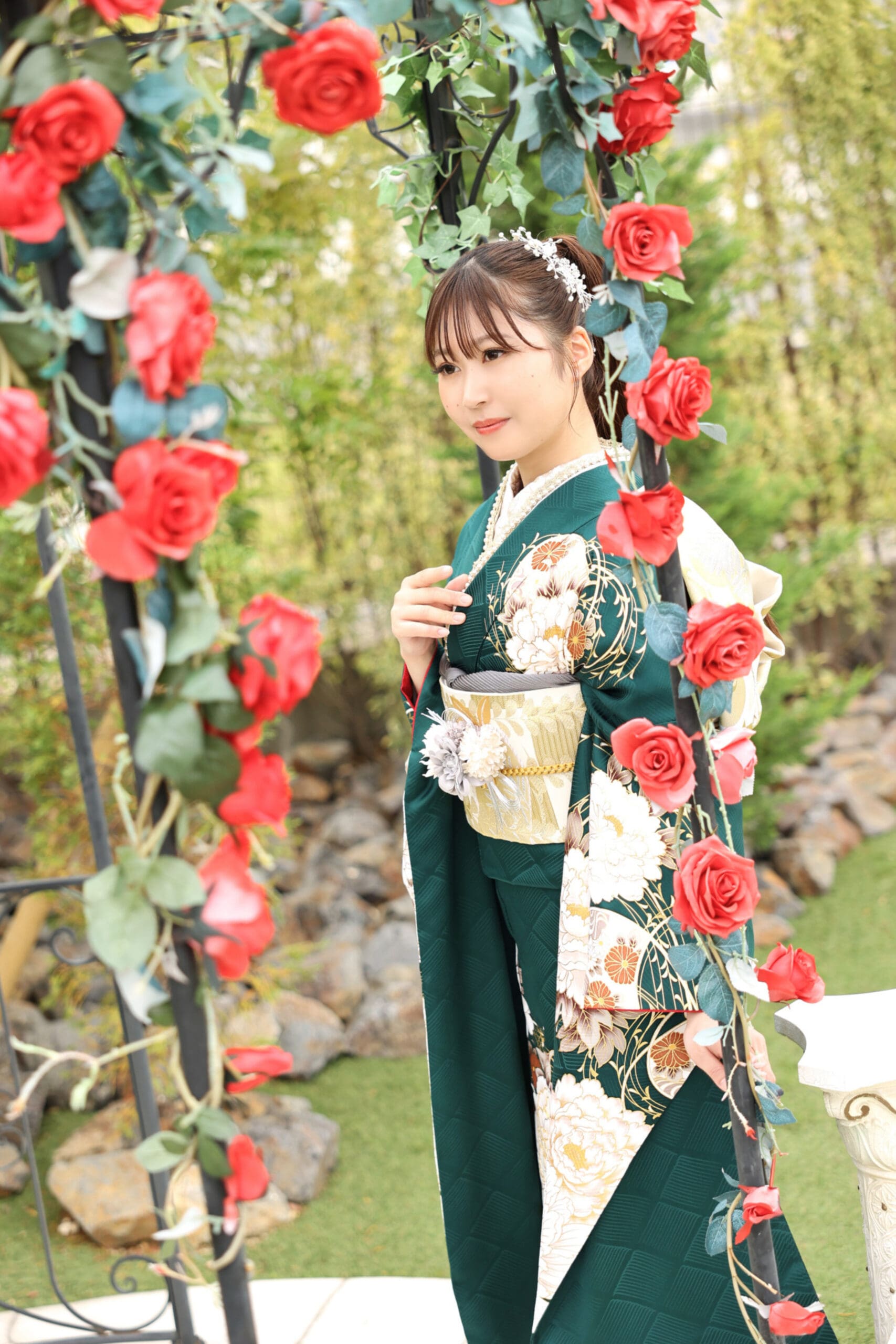 鮮やかな赤いバラで飾られたアーチに縁取られた庭に、花模様の濃い緑の着物を着た若い女性が立っている。花柄のヘアアクセサリーをつけた彼女は、緑と岩を背景に柔らかな微笑みを浮かべている。.
