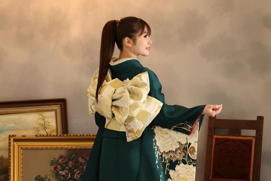 長い茶髪をポニーテールにした女性が、花模様の緑色の着物を着て、金と白の大きな飾り帯を締めている。額装された美術品と木製の椅子が背後に置かれた柔らかな照明の部屋で、彼女は横向きに微笑みながら立っている。.