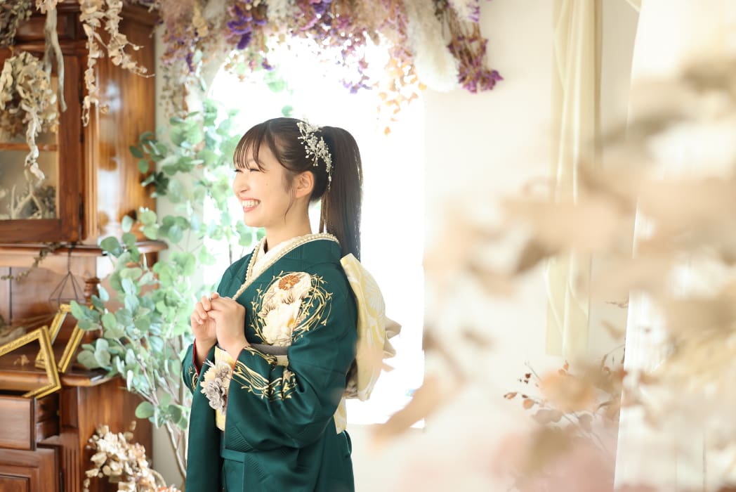 花柄の刺繍が施された緑色の着物を着た若い女性が、室内で手を合わせて微笑んでいる。彼女は髪飾りをつけ、ドライフラワーと緑に囲まれている。.