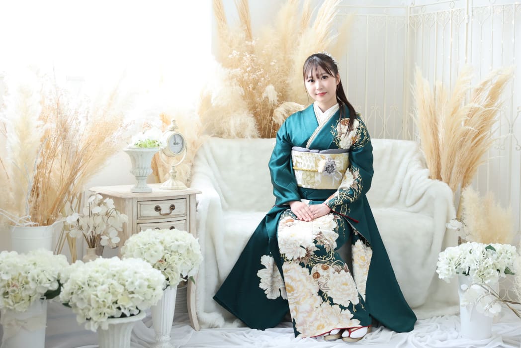 緑とクリーム色の花柄の着物を着た若い女性が白いソファに座っている。白い花とベージュのパンパスグラスで飾られた明るくエレガントな空間は、柔らかく落ち着いた雰囲気を醸し出している。優しく微笑み、髪飾りをつけたストレートの黒髪。.