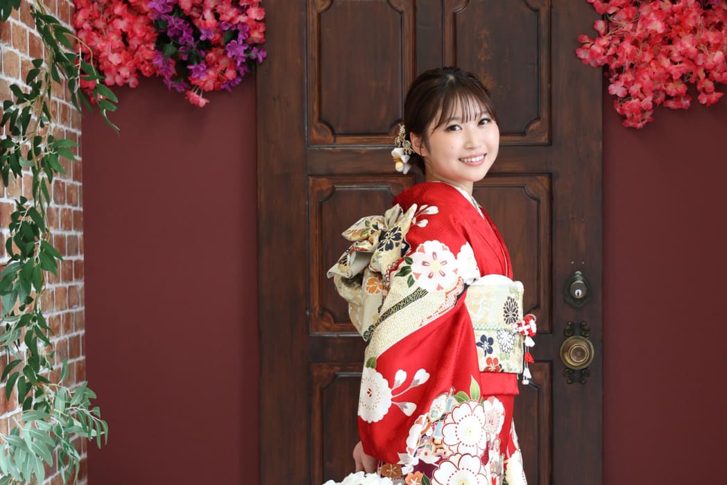 赤と白の花柄の着物を着た女性がカメラに向かって微笑み、金属製の装飾が施された木製のドアの前に立っている。上部の角にはピンクと紫の花が飾られ、レンガの壁の左側には緑の葉がぶら下がっている。.