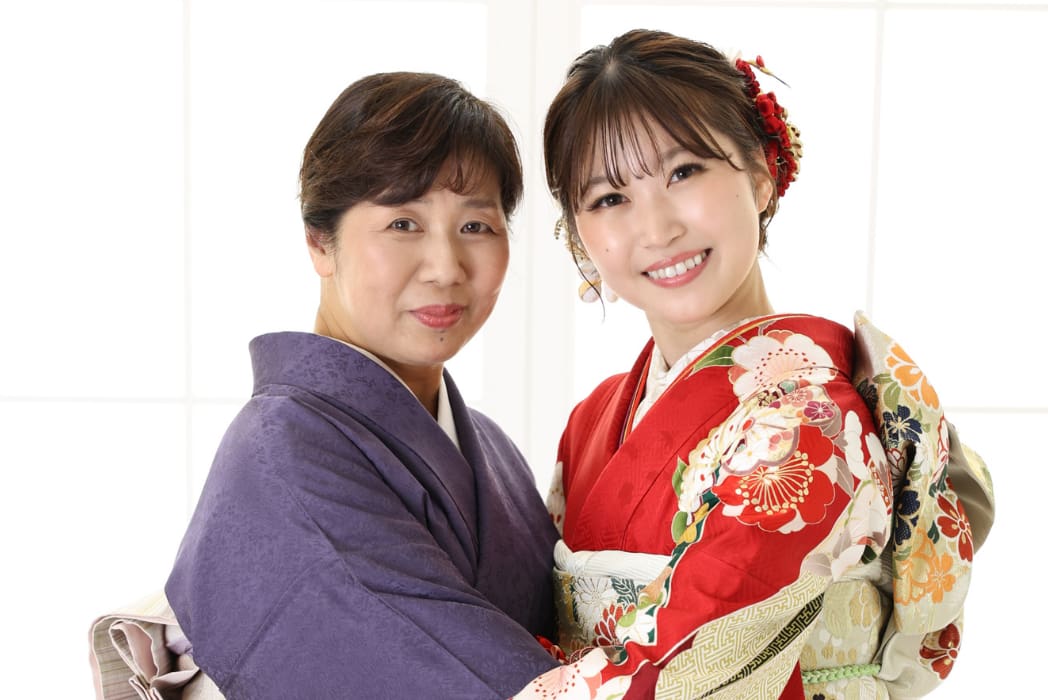 赤い花柄の着物を着た笑顔の若い女性が、紫の着物を着た年配の女性の隣に立っている。ふたりはカメラに向かい、寄り添い、抱き合い、背後には窓から差し込む明るい自然光がある。.