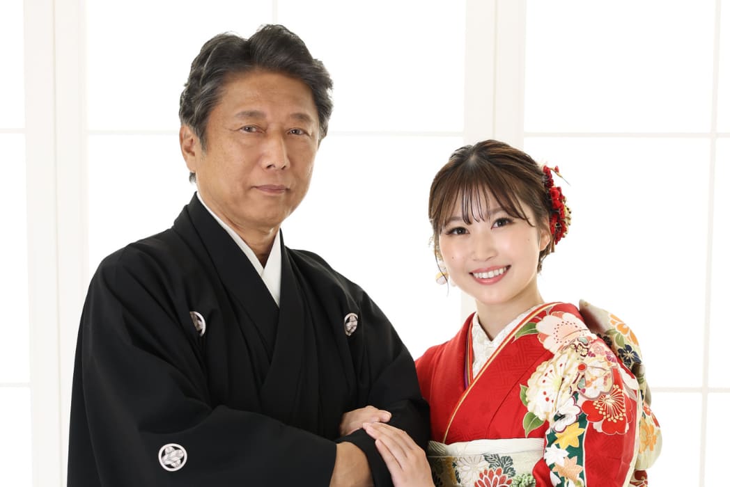 明るい窓の前で、花模様の鮮やかな赤い着物を着て髪飾りをつけた若い女性が笑顔でポーズをとっている横に、日本の伝統的な黒い着物を着た男性が立っている。二人は楽しそうで仲が良さそうだ。.