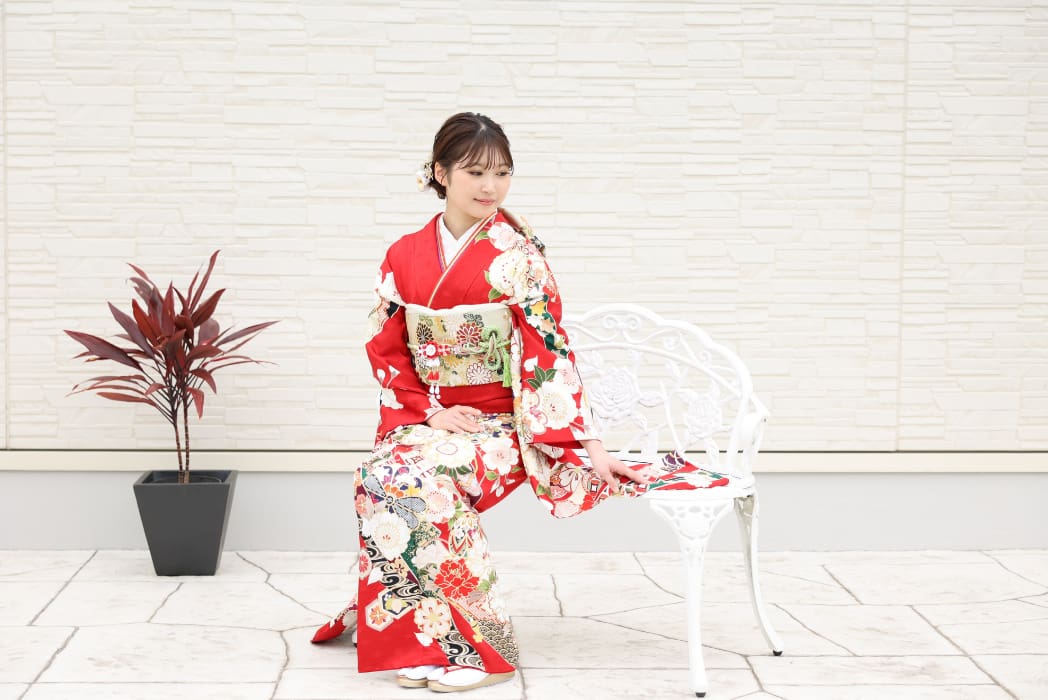 鮮やかな赤い花柄の着物を着た若い女性が、白い金属製のベンチに優雅に座っている。髪に花を飾り、着物の袖を押さえて下を向いている。彼女の左側には赤い葉をつけた鉢植えが、淡い石壁を背にして置かれている。.