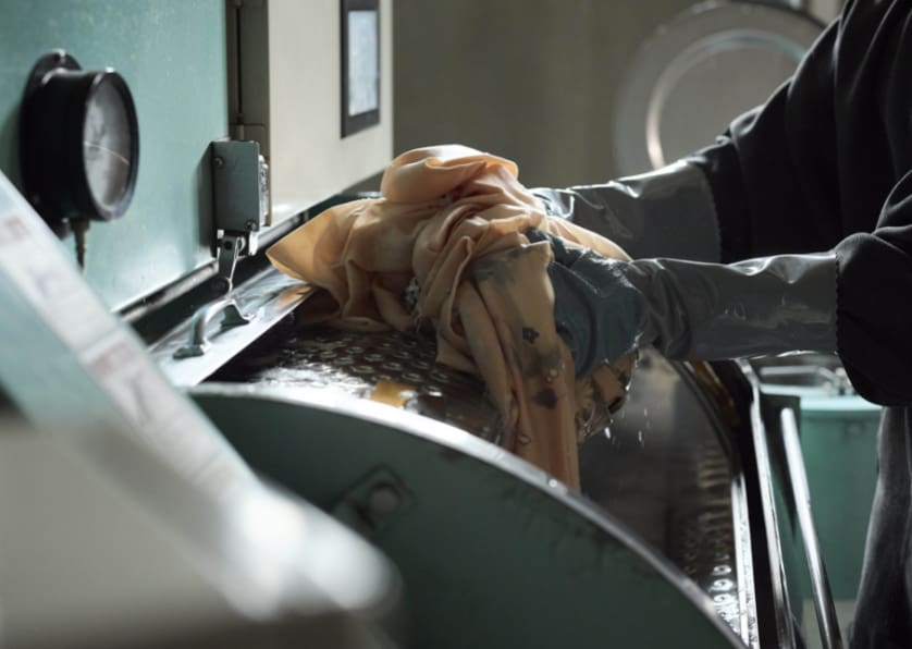 灰色のゴム手袋をはめた人物が、大きな工業用洗濯機に桃色の布地を投入している。洗濯機は金属製で、ダイヤルが見え、蓋が開いている。背景はぼやけ、部分的に光が当たっている。.