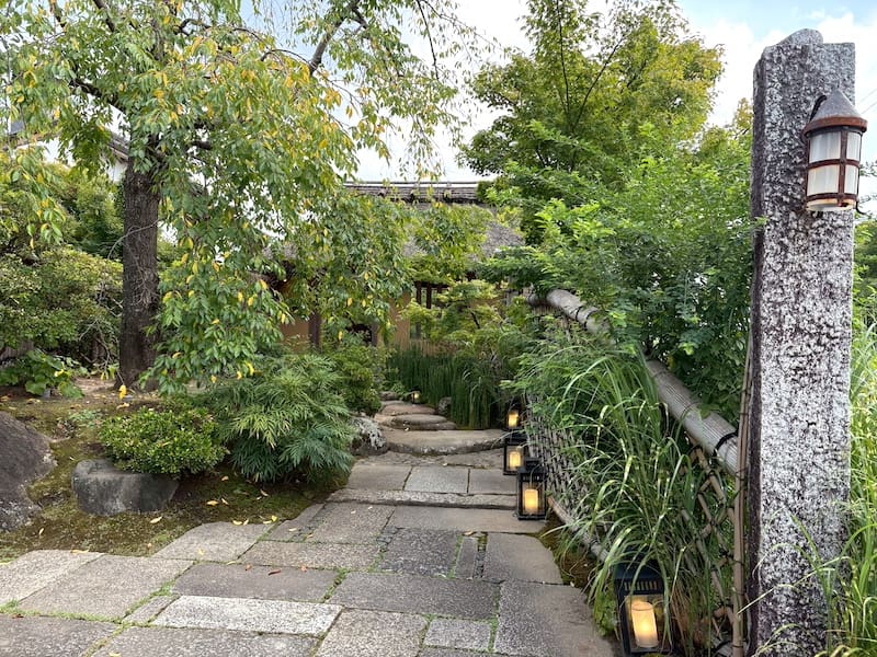 灯籠が並ぶ石畳の小道は、緑豊かな木々の間を通り、伝統的な木造の日本建築へと続く。風化した石柱と竹垣が、着物の散歩やもみじ狩りに最適な落ち着いた庭の風景をさらに引き立てている。.