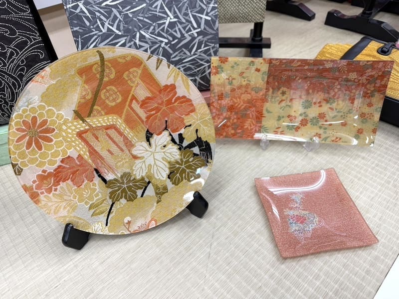 着なくなった着物をイメージした複雑な和風の花模様が描かれた丸い飾り皿、長方形の盆、四角い小皿が、ベージュの質感の表面に、他の模様の小物を背景に飾られている。.