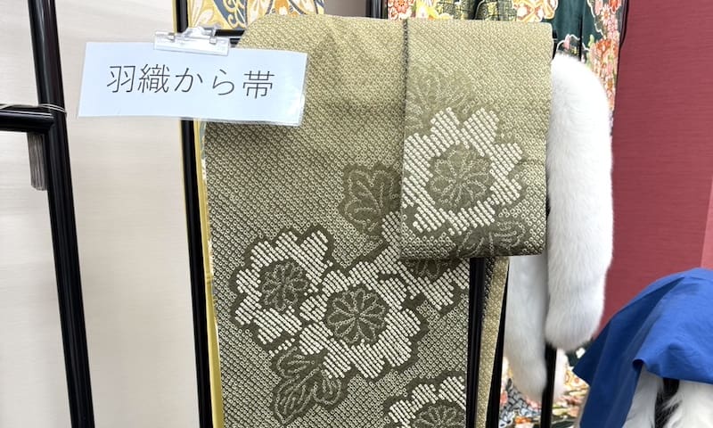 緑と金の花模様の飾り帯が飾られている。その上には羽織から帯と書かれた白い看板が掲げられている。背景には着物やスタッフブログ用の伝統的な衣服が飾られている。.