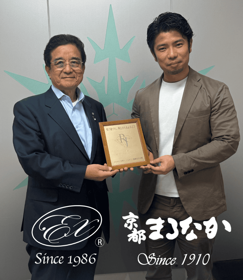 2人の男性が並んで立ち、笑顔で一緒にプラークを掲げています。1人はメガネをかけ、ダークスーツに青いシャツを着ており、もう1人は白いシャツの上にベージュのスーツを着ています。 二人の背後には、下部に日本語と英語のテキストが記載されたスタイリッシュなロゴがあり、この場面が「ジュエリー相談会」に関連しているか、あるいは「スタッフブログ」で取り上げられていることを示唆している。.