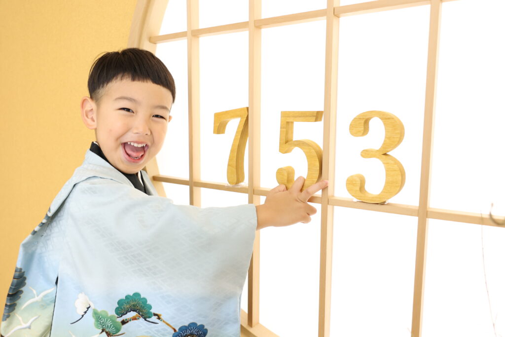 水色の柄入りの着物を着た少年が、障子風の屏風に貼られた木製の数字753を指さしながら笑っている。その光景は明るく照らされ、柔らかくミニマルな美しさを醸し出している。.